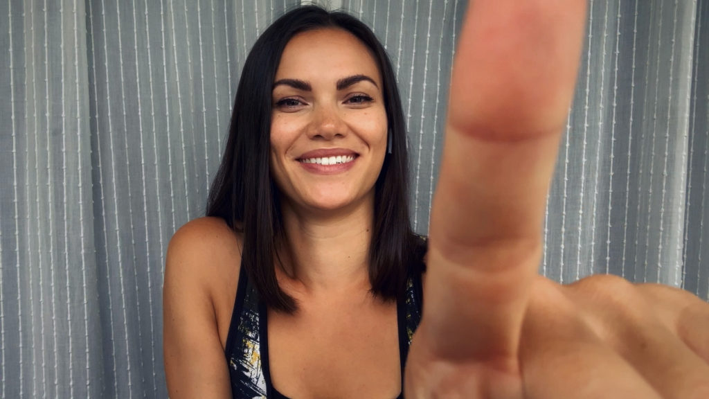 Screenshot. Szene mit Paige vor einem grauen Vorhang, lächelnd und Kamera mit einem Finger ausschaltend.