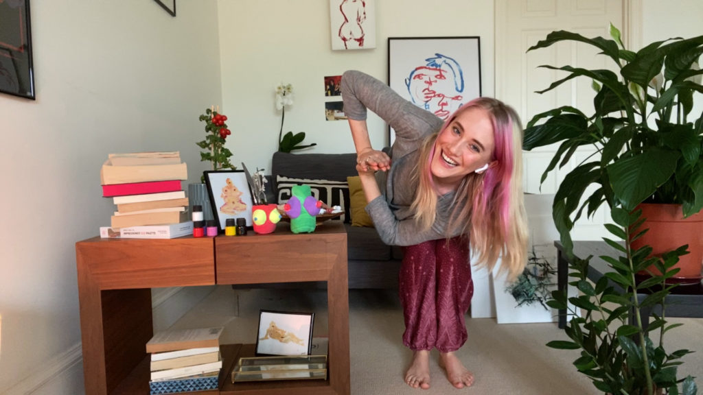 Screenshot. Szene mit Saffron, die vor der Kamera eine Yoga-Pose macht. Links neben ihr steht ein Tisch mit Büchern sowie Kunstskulpturen.