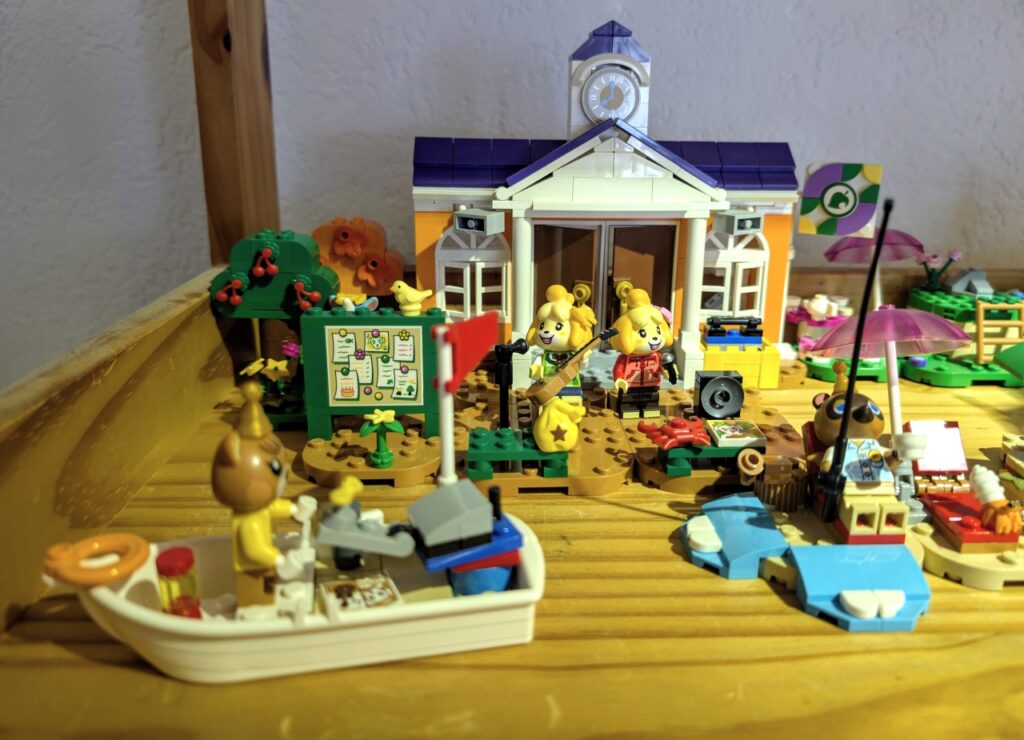 Blick auf das Rathaus. Kleines Boot im Vordergrund, dahinter Schlagbrett. Melinda und Melinda an Gitarre und Mikrofon stehen vor der Rathaustür. Rechts neben dem Boot ist ein wenig Wasser zu sehen und Tom Nook auf einer Strandliege mit Angel in der Hand