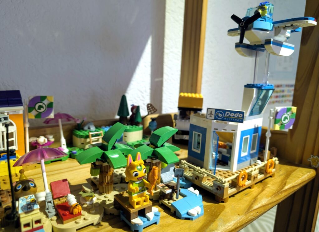 Strand und Flughafen. Das Flugzeug ist mit einem durchsichtigen Legostück über dem Flughafen befestigt. Auf einem schmalen Steg sitzt Tanja mit einem Fisch.