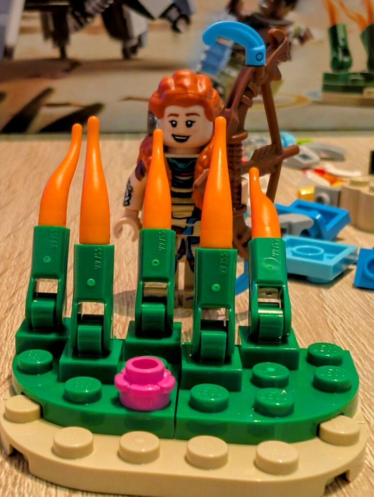 Aloy steht hinter dem Lego-Gras und lächelt.