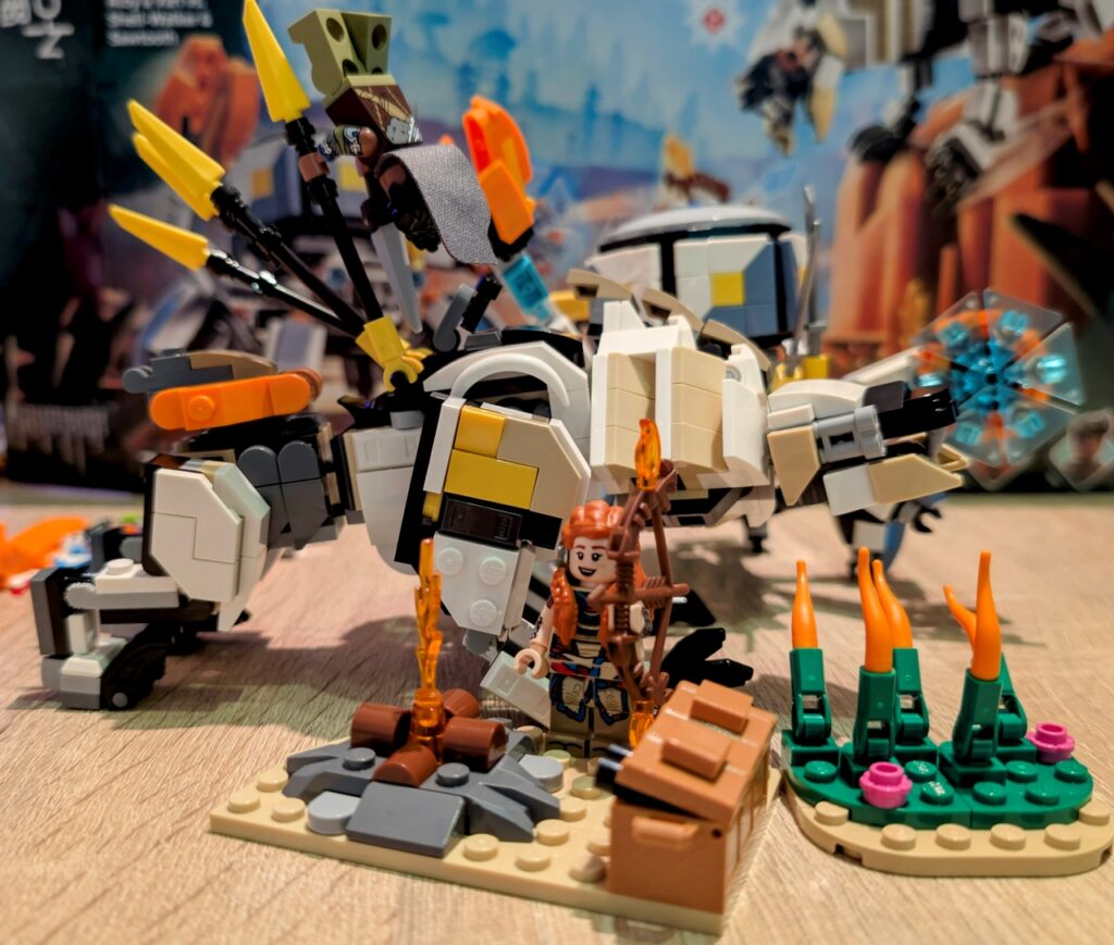 Das gesamte Set zusammengedrängt auf einem Foto. Vorne Lagerfeuer, Gras und lächelnde Aloy mit Bogen, dahinter Varl kopfüber an einer Antenne des Sägezahns befestigt. Halb versteckt dahinter der Panzerwanderer.