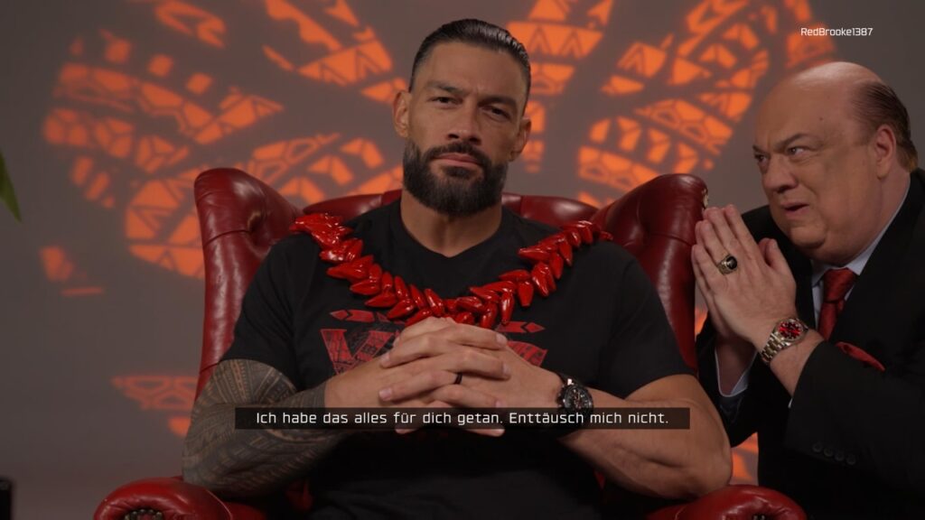 Nahaufnahme des echten Roman Reigns auf rotem Ledersessel, daneben Paul Heynes mit aufeinanderliegenden Handflächen. Reigns: "Ich habe das alles für dich getan. Enttäusch mich nicht."