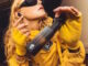 Foto einer weiblichen Person in gelben Ganzkörperanzug mit Sonnenbrille. Sie hält das Case der Method 360 ANC in den Fokus der Kamera und hölt den Kopf schief, um ein In-Ear zu sehen.
