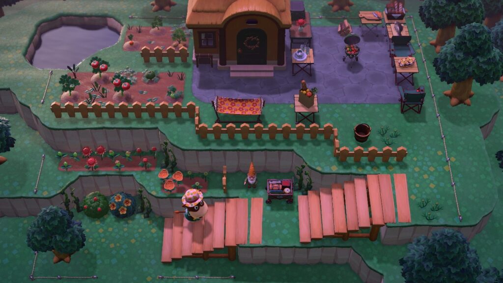 Blick von oben auf das Grill-Lysium. Links ein Teich, daneben zwei Felder, eingerahmt von Zäunen. Deneben ist das Haus mit einem Türkranz aus Zweigen. Rechts davon der Grillplatz mit mehreren Klapptischen aus Holz, darauf Geschirr, Schneidebrett, Brotkorb, Fisch und Grillkartoffeln. Vorne die beiden Holztreppen als Aufgänge. Auf dem ersten Absatz steht ein kleiner Handwagen.