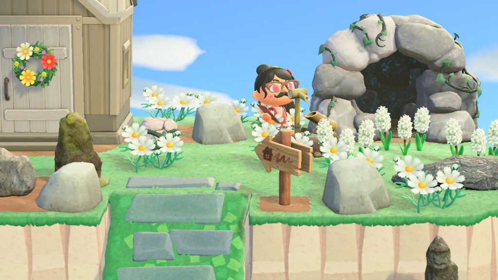 Screenshot vom Ort zum Bergsteigen. Nahaufnahme der oberen Ebene mit Spielfigur in der Mitte, links Haus, rechts Höhle, überall Blumen, Steine und Felsen. Vor der Spielfigur das Wasserspiel.