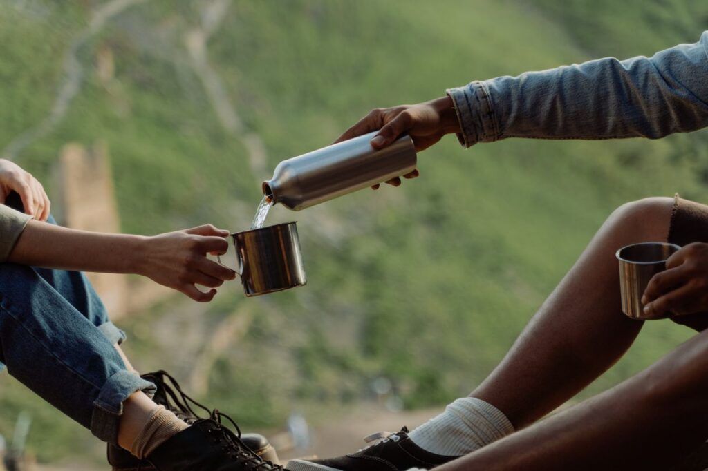 Foto von zwei Menschen. Die rechte Person schüttet der Person links Wasser aus einer Flasche in eine Tasse.